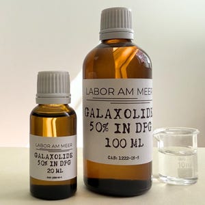 May include: Two amber glass bottles with white caps and labels reading Labor Am Meer Galaxolide 50% in DPG, one 20 ml and one 100 ml, alongside a small clear glass beaker with 10 ml markings, containing a clear liquid, all on a light surface.
