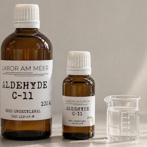 Puede incluir: Dos botellas de vidrio marrón y un vaso de precipitados pequeño. Las botellas tienen etiquetas blancas con texto negro que dice "LABOR AM MEER ALDEHYDE C-11". Una botella está etiquetada 100 ml, la otra 20 ml. El vaso contiene un líquido transparente.