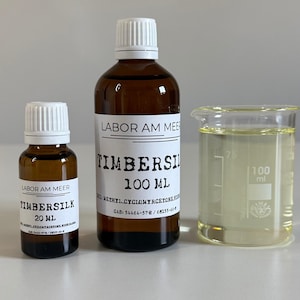 May include: Three amber glass bottles and a beaker. The bottles are labelled "TIMBERSILK" with volumes of 20 ml and 100 ml. The beaker contains a yellow liquid and has 100 ml markings. The text "LABOR AM MEER" is also visible.