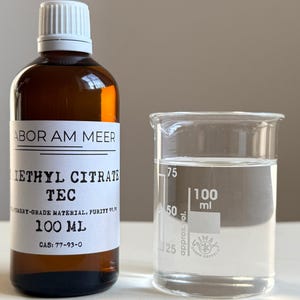May include: A brown glass bottle with a white label and a white cap, next to a clear glass beaker. The bottle label reads "IETHYL CITRATE TEC" and "100 ml". The beaker contains a clear liquid, with 100 ml markings. The purity is 99.9%.