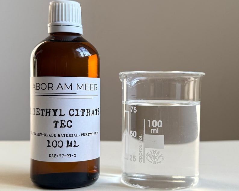 May include: A brown glass bottle with a white label and a white cap, next to a clear glass beaker. The bottle label reads "IETHYL CITRATE TEC" and "100 ml". The beaker contains a clear liquid, with 100 ml markings. The purity is 99.9%.