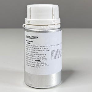 Może przedstawiać: Srebrna, cylindryczna butelka z białą zakrętką. Butelka posiada białą etykietę z czarnym tekstem, w tym "ISO E SUPER CRYSTAL" i inne informacje chemiczne. Butelka zawiera 120 ml płynu.