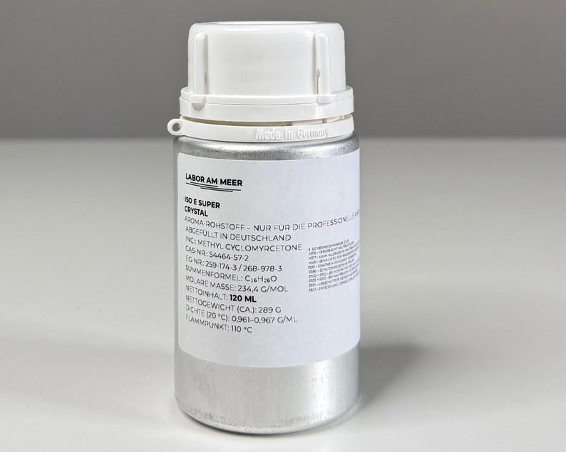 Może przedstawiać: Srebrna, cylindryczna butelka z białą zakrętką. Butelka posiada białą etykietę z czarnym tekstem, w tym "ISO E SUPER CRYSTAL" i inne informacje chemiczne. Butelka zawiera 120 ml płynu.