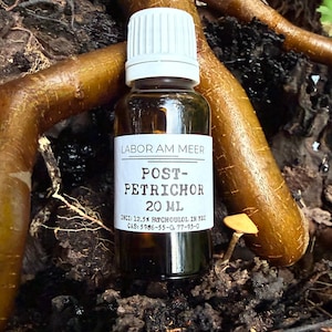 Può includere: Una piccola bottiglia di vetro marrone con tappo bianco, etichettata "POST-PETRICHOR 20 ml." La bottiglia è appoggiata su terreno scuro con radici esposte e un piccolo fungo. L'etichetta include il testo "LABOR AM MEER."