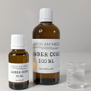 May include: Three amber glass bottles with white caps and labels. The labels read "LABOR AM MEER AMBER CORE" with sizes 20 ml and 100 ml. A small beaker with liquid is also present.