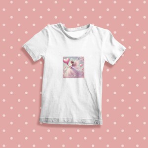 K&ouml;nnte beinhalten: Wei&szlig;es T-Shirt mit einem Motiv einer Fee, die einen herzf&ouml;rmigen Ballon h&auml;lt. Die Fee tr&auml;gt ein wei&szlig;es Kleid und hat rosa Fl&uuml;gel. Der Hintergrund ist hellrosa mit wei&szlig;en Punkten.