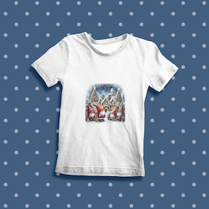 Puede incluir: Camiseta blanca con una escena navide&ntilde;a con tres gnomos de pie frente a una casa cubierta de nieve con un &aacute;rbol de Navidad. Los gnomos llevan gorros rojos y tienen barbas blancas.