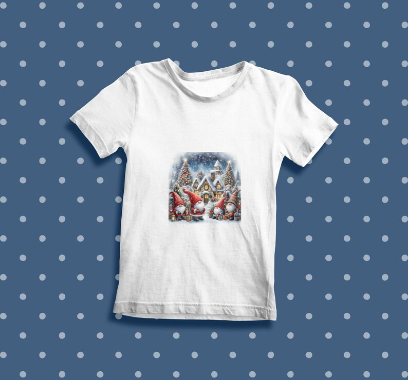 Puede incluir: Camiseta blanca con una escena navide&ntilde;a con tres gnomos de pie frente a una casa cubierta de nieve con un &aacute;rbol de Navidad. Los gnomos llevan gorros rojos y tienen barbas blancas.