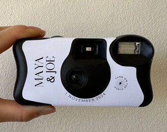 Puede incluir: Una cámara desechable negra con una funda blanca personalizada que presenta el texto MAYA & JOE, la fecha 1 NOVEMBER 2024 y un icono circular con el texto TURN ON THE FLASH. La cámara incluye una lente central, un visor y un flash rectangular.