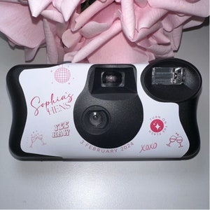 May include: Black and white disposable camera with a white label. The label has the text "Sophia's HENS", "Yee Haw", and "3 February 2024". The camera has a flash and a lens. A pink rose is in the background.