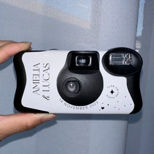 May include: White disposable camera with black accents. The camera has the text "Amelia & Lucas" and "14 November 2023" printed on it. The camera also has a black circle with the text "FLASH" and "TURN ON THE FLASH" printed on it.