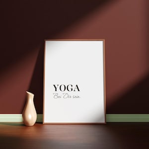 Peut inclure: Une affiche encadrée avec le mot "YOGA" et la phrase "Bei Dir sein." en noir. Un vase beige est placé à gauche de l'affiche. Le fond est un mur rouge foncé avec un sol en bois.