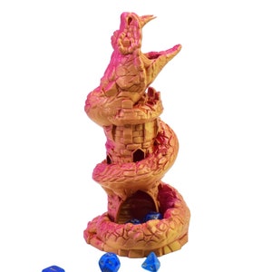 Puede incluir: Una torre de dados de fantasía en oro y rosa. La torre tiene un diseño en espiral con una estructura similar a un castillo y una boca abierta en la parte superior. Varios dados azules están esparcidos por la base.