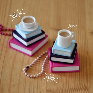 Può includere: Due ciondoli in miniatura a forma di pile di tre libri con sopra una tazzina da caffè bianca. Ogni pila presenta un libro azzurro, uno blu scuro e uno rosa. Una catenella a palline rosa è attaccata a una pila, una rosa chiaro all'altra.