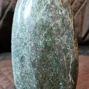 May include: An oval-shaped stone sculpture with a smooth, polished surface. The object features a deep forest green color with mottled textures, subtle brown mineral inclusions, and a reflective sheen across its rounded, elongated vertical form.
