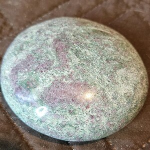 May include: A smooth, polished, oval-shaped stone featuring a mottled pattern of deep forest green and vibrant magenta-purple mineral inclusions. The surface has a glossy, reflective finish that highlights the natural texture and color variations.