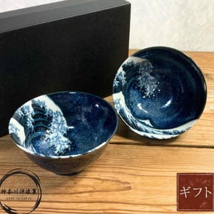 Puede incluir: Dos cuencos de cerámica azules y blancos con un diseño de olas. Los cuencos están sobre una superficie de madera, con una caja negra en el fondo. Los cuencos tienen un acabado brillante y un interior azul oscuro.