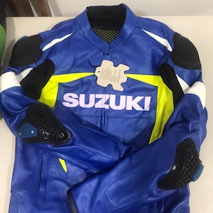 May include: A blue leather motorcycle jacket with yellow and white accents. The jacket has the word "SUZUKI" printed on the front in white letters. The jacket has protective padding on the elbows and shoulders.