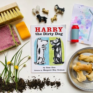 May include: A children's book titled "Harry the Dirty Dog" by Gene Zion with illustrations by Margaret Bloy Graham. The book cover features a black and white dog with a red collar and a white dog with a blue collar. The book is surrounded by various items including a brush, a bar of soap, a pink cloth, a bottle of bubble solution, a straw, a plate of dog biscuits, and a small pile of dirt.
