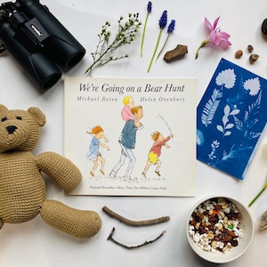 Könnte beinhalten: Ein brauner gehäkelter Teddybär, ein Buch mit dem Titel "We're Going on a Bear Hunt" von Michael Rosen und Helen Oxenbury, ein blauer Cyanotypieabdruck, eine Schüssel mit Studentenfutter, ein Fernglas und verschiedene Naturgegenstände wie Stöcke, Blumen und Steine.
