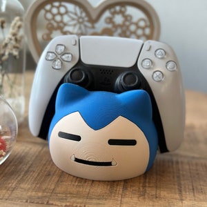 Könnte beinhalten: Ein weißer PlayStation 5-Controller wird in einem blau-weißen 3D-gedruckten Ständer gehalten, der die Form eines Pokémon-Charakters hat. Der Ständer hat ein weißes Gesicht mit schwarzen Augen und einem kleinen Lächeln.