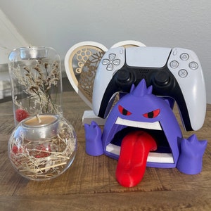 Könnte beinhalten: Lila und roter 3D-gedruckter Videospiel-Controller-Ständer in Form eines Cartoon-Monsters mit einem großen Maul und einer Zunge. Der Ständer hält einen weißen drahtlosen Videospiel-Controller.