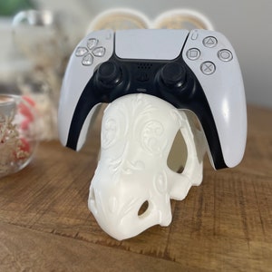Könnte beinhalten: Ein weißer PlayStation 5 DualSense-Controller liegt auf einem weißen, 3D-gedruckten Schädel-Ständer. Der Controller hat schwarze Akzente und Knöpfe.