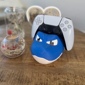 Könnte beinhalten: Ein blauer und weißer Videospiel-Controller-Ständer in Form einer Comicfigur mit wütenden Augenbrauen. Der Ständer hält einen weißen PlayStation 5-Controller.