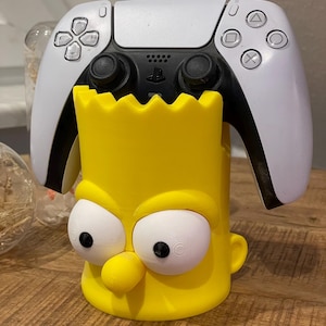 Könnte beinhalten: Ein gelber Controller-Halter im Bart Simpson-Design mit großen weißen Augen und einer markanten Nase. Ein weißer und schwarzer PlayStation 5-Controller ruht in der Halterung. Der Halter ist aus Kunststoff gefertigt und für einen Gamecontroller konzipiert.