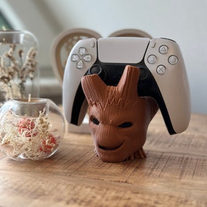 Könnte beinhalten: Ein brauner, Groot-förmiger Videospiel-Controller-Ständer mit einem weißen PlayStation 5-Controller, der darauf liegt.