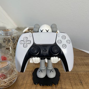 Könnte beinhalten: Ein weißer und grauer PlayStation-Controller-Ständer mit einer stilisierten Figur mit einem 'X'-Design. Der Ständer hält einen weißen PlayStation 5-Controller.