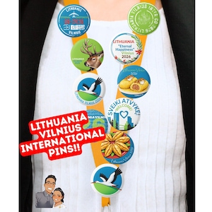 Puede incluir: Una colección de coloridos pines de Lituania Vilnius International. Los pines presentan varios diseños, incluyendo un ciervo, una cigüeña y el texto "Eternal Happiness Vilnius 2026". Los pines están sujetos a un cordón naranja.