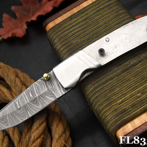 May include: A silver pocket knife with a patterned blade and a brass accent. The knife is closed and resting on a wooden box with a green and brown striped fabric covering.