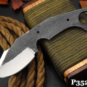 May include: A dark gray, unfinished knife blade with a polished silver edge. The blade has a unique shape with a circular hole near the handle. The knife rests on a wooden and green striped sheath, with a brown rope in the background.