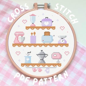 May include: A round wooden embroidery hoop containing a cross stitch pattern of kitchen appliances on three shelves. The design features a blender, pot, mixer, toaster, scale, coffee grinder, and moka pot in pastel colors with small heart motifs.