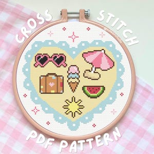 May include: A cross stitch pattern in a round wooden hoop featuring a heart-shaped design with a light blue scalloped border. The interior displays summer-themed icons including pink heart sunglasses, a suitcase, an ice cream cone, a pink beach umbrella, a watermelon slice, and a yellow sun on a pale yellow background.