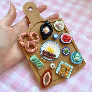 May include: A wooden cutting board with a variety of miniature food items, including pretzels, a sausage skewer, a fried egg, a small bowl of peas, a small bowl of cucumbers, a small bowl of red sauce, a small bowl of rice, a small bowl of white sauce, a small bowl of meatballs, and a small bowl of pastries. The cutting board is decorated with a green and white checkered pattern.