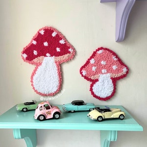Peut inclure: Deux décorations murales en forme de champignon rose et blanc avec des pois blancs. Les champignons sont faits de laine et sont accrochés à un mur blanc. Il y a quatre voitures miniatures sur une étagère bleu clair sous les champignons.