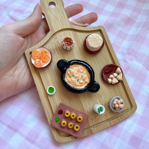 May include: Miniature food items arranged on a small wooden cutting board. The food includes a small pot of stew, a stack of pancakes, and other dishes. The board is held by a hand, and the background is a pink and white checkered pattern.