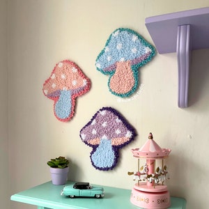 May include: Three colorful mushroom-shaped wall decorations made of yarn. The mushrooms are in shades of pink, purple, and blue with white polka dots. The mushrooms are hanging on a white wall with a purple shelf above them.