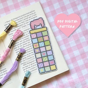 Könnte beinhalten: Ein Kreuzstich-Lesezeichen mit einem Gitter aus pastellfarbenen Quadraten in Gelb, Rosa, Lila und Blau, gekrönt von einem rosa Katzenohren-Design. Der Text PDF DIGITAL PATTERN steht in einem rosa Herz. Vier Stickgarnstränge sind abgebildet.