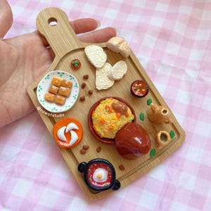 May include: A wooden cutting board with a variety of miniature food items, including bread, couscous, a tagine, and a bowl of orange squares. The board is decorated with green leaves and brown beans.