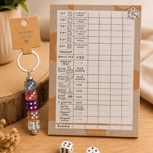 Könnte beinhalten: Ein Schlüsselanhänger mit fünf durchsichtigen, bunten Würfelperlen an einem Metallring mit einem braunen Anhänger mit der Aufschrift Handmade with love. Daneben liegt ein Spielblock mit deutschem Text für ein Würfelspiel und drei weiße Würfel.