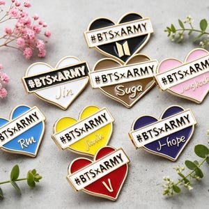 Peut inclure: Collection de broches en émail en forme de cœur de différentes couleurs, chacune avec le texte "#BTSxARMY" et un nom. Les broches ont des bordures dorées et sont disposées avec de la verdure et des fleurs décoratives.