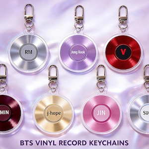 May include: A collection of seven vinyl record-shaped keychains in various colors, each featuring a different name or initial. The keychains have silver-colored metal clasps and are displayed against a soft, light purple background. The text "BTS VINYL RECORD KEYCHAINS" is at the bottom.