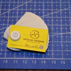 May include: A yellow card with the text "OFFICIAL LOGBOOK" and a compass logo. The card is printed on "Rite in the Rain" all-weather writing paper. The card is sitting on a blue gridded surface.