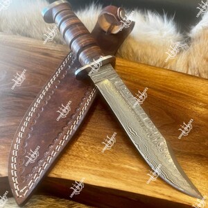 May include: A Damascus steel hunting knife with a brown leather handle and sheath. The blade has a unique patterned design. The sheath is also brown leather with white stitching. The knife is displayed on a wooden surface.