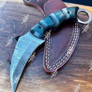 May include: A karambit knife with a curved blade and a finger ring. The handle is a mix of blue and black, and the blade has a unique pattern. It rests on a brown leather sheath and a wooden surface.