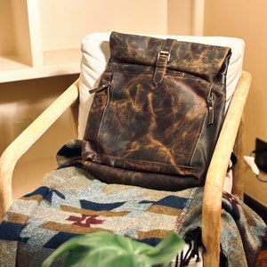 May include: A brown leather backpack with a flap closure and a buckle. The backpack is sitting on a chair with a patterned blanket.