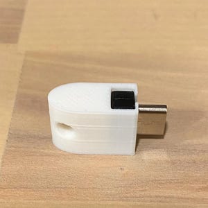 May include: A white USB adapter with a black square button and a silver USB connector. The adapter is on a light brown wooden surface. The adapter is a small electronic device.